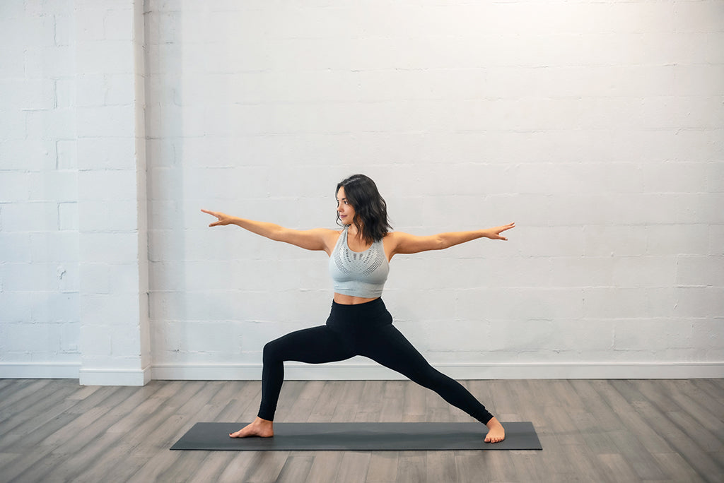 A person stands on a yoga mat in a well-lit room, performing the Warrior II pose. They are wearing a grey sports bra and black leggings, extending their arms parallel to the floor with one leg bent forward and the other straight behind them.