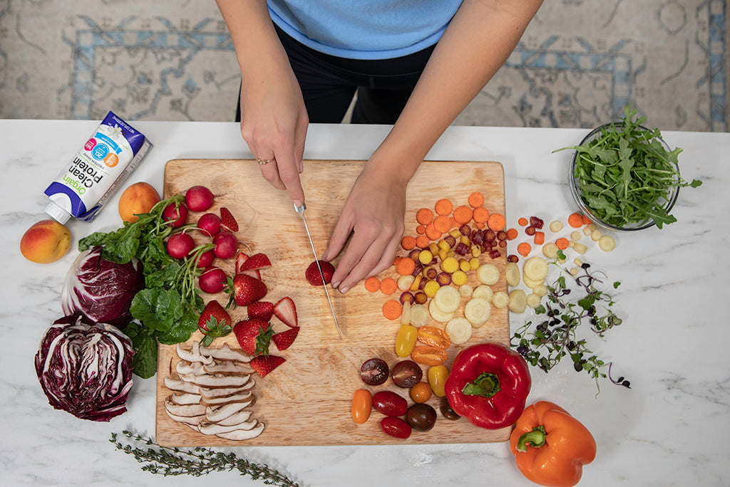 A person is chopping a strawberry on a wooden cutting board. Surrounding the board are various fresh vegetables and fruits, including radishes, carrots, bell peppers, mushrooms, leafy greens, and a box of protein blend drink.
