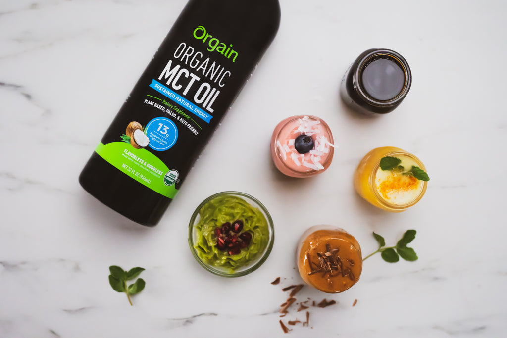A bottle of Orgain Organic MCT Oil lays on a marble surface alongside five small glass jars containing various food items with different colors and textures. The jars are garnished with items like pomegranate seeds, chocolate shavings, and herbs.