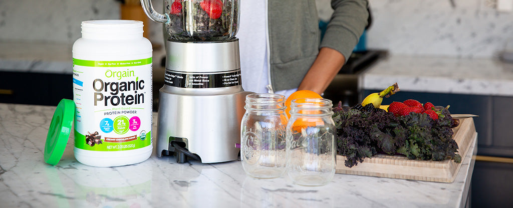 A person prepares a smoothie in a blender with fresh strawberries visible inside. On the counter are a container of Orgain Organic Protein powder, two mason jars, leafy greens, an orange, a banana, and strawberries on a cutting board.