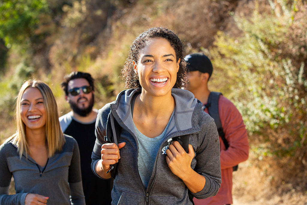 Four hikers on a sunny trail, focusing on a smiling woman in a gray jacket, with a wooded area behind.