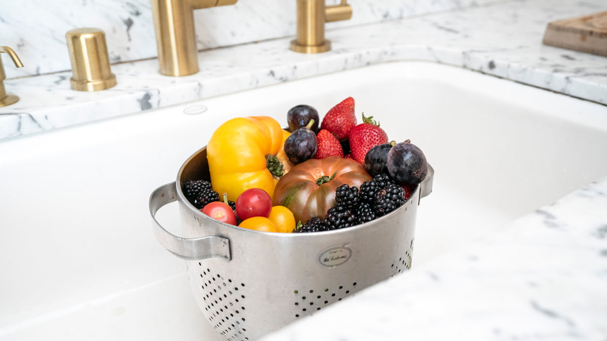 A metal colander filled with assorted fresh produce, including yellow bell peppers, a tomato, strawberries, blackberries, and small plums, sits in a white marble sink. The kitchen's gold-colored faucet and fixtures are visible in the background.