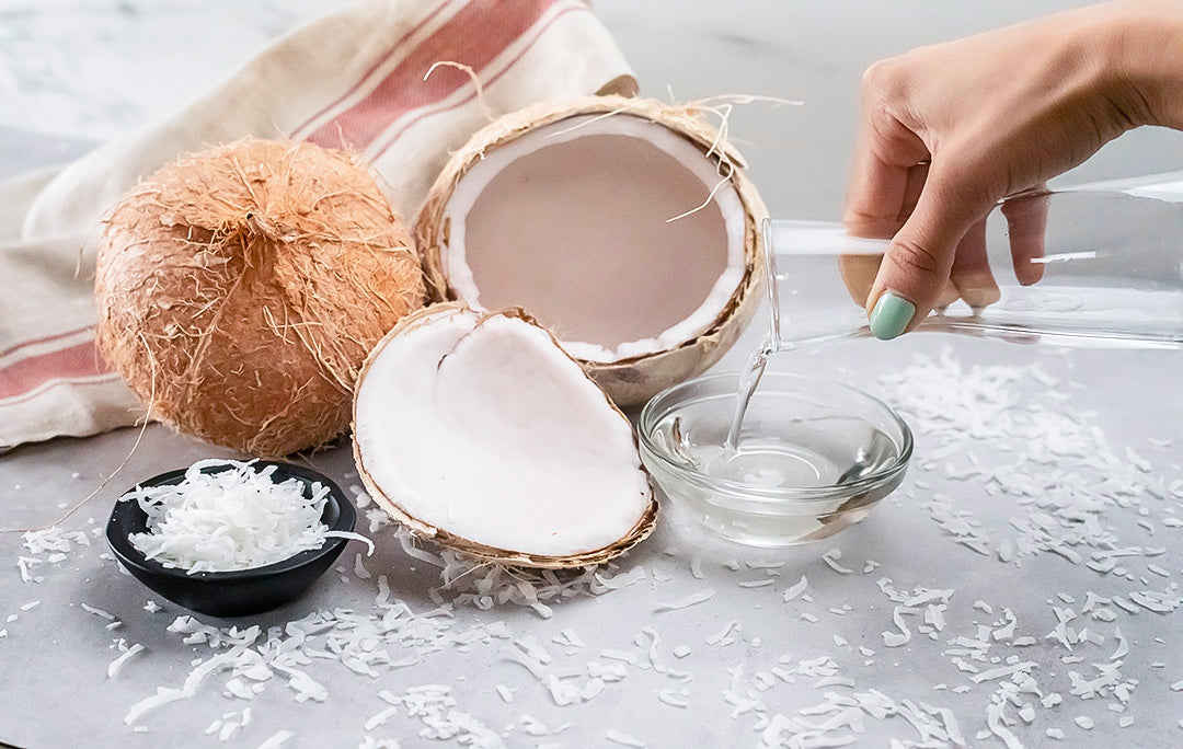 A person's hand pouring coconut water into a small glass bowl, surrounded by a whole coconut, a cracked coconut, shredded coconut in a black bowl, and loose shredded coconut on a flat surface with a red-striped cloth in the background.