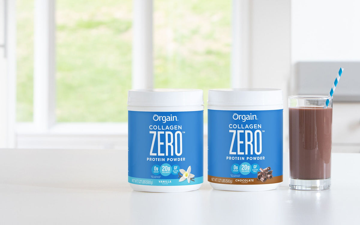 Two containers of Orgain Collagen Zero Protein Powder (Vanilla and Chocolate flavors) on a white surface with a glass filled with a chocolate-colored drink and a blue-striped straw.