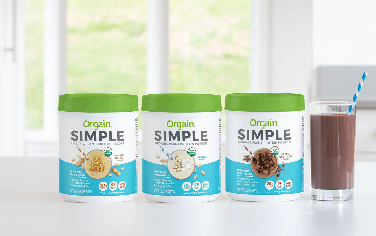 Three containers of Orgain Simple Organic Plant Protein Powder in Vanilla, Peanut Butter, and Creamy Chocolate flavors are displayed on a white surface. A glass of chocolate protein shake with a blue and white striped straw is positioned to the right.
