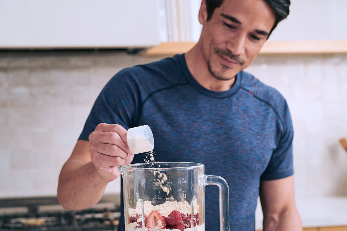 A man in a blue shirt is making a smoothie in a kitchen. He is pouring a scoop of protein powder into a blender filled with ingredients like strawberries and oats. The kitchen counter and backsplash are visible in the background.
