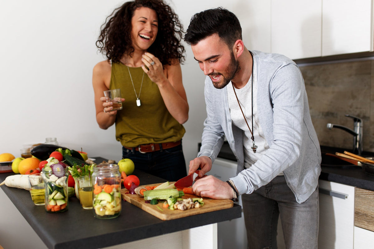 A man and woman are in a modern kitchen. The man is chopping vegetables on a wooden cutting board, while the woman stands nearby, holding a glass and laughing. The counter is covered with various fresh vegetables, fruit, and jars of colorful salads.