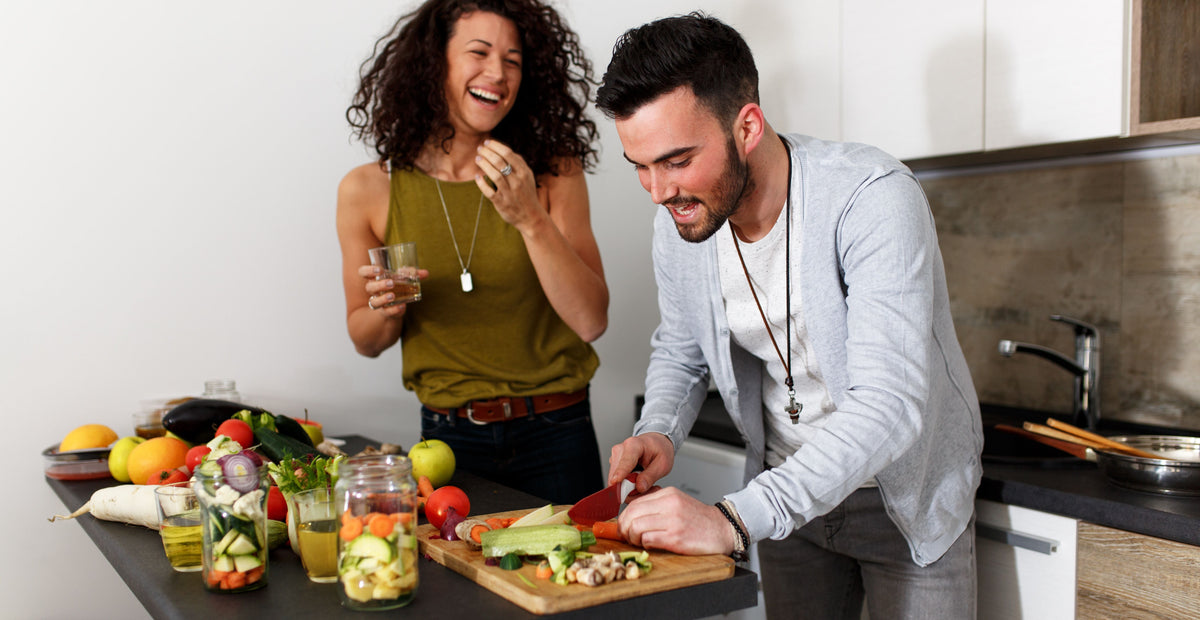 A woman and a man are in a kitchen. The woman, holding a drink, is laughing, while the man is cutting vegetables on a chopping board. Various fruits and jars of chopped vegetables are on the counter. They appear to be enjoying their time cooking together.