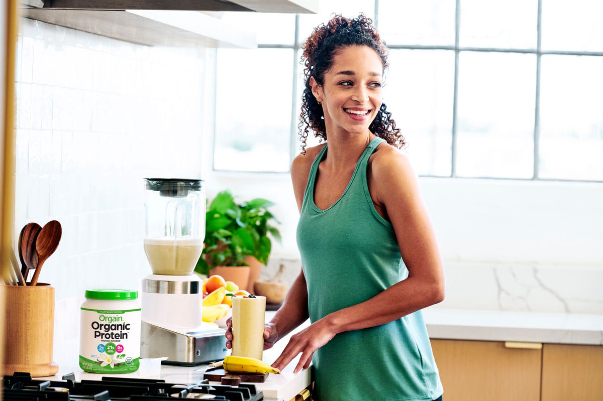 A woman in a green tank top stands in a bright kitchen. She is smiling and holding a glass of a smoothie. A blender and a container of Orgain Organic Protein powder are on the counter, along with a banana and a bowl of fruit.