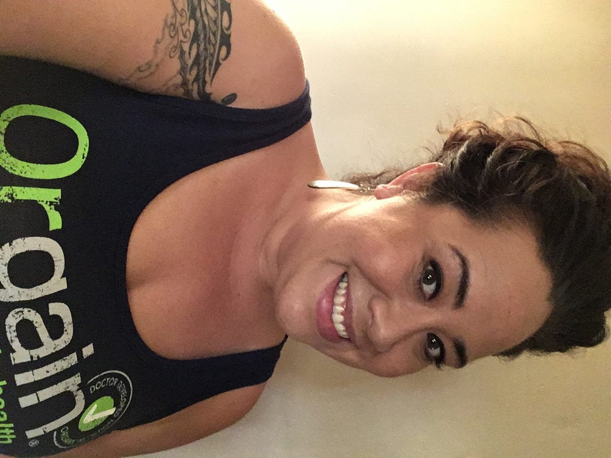 A woman with curly dark hair tied up, wearing a navy blue tank top with visible printed text, smiles at the camera. She has a tattoo on her upper arm and stands against a neutral background.