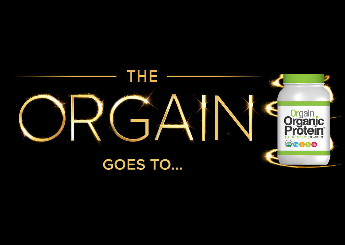 An image with the text "The ORGAIN goes to..." in bright, glowing gold letters. To the right, there is a container of Orgain Organic Protein plant-based powder, featuring the brand's logo and label on the bottle. The background is black.