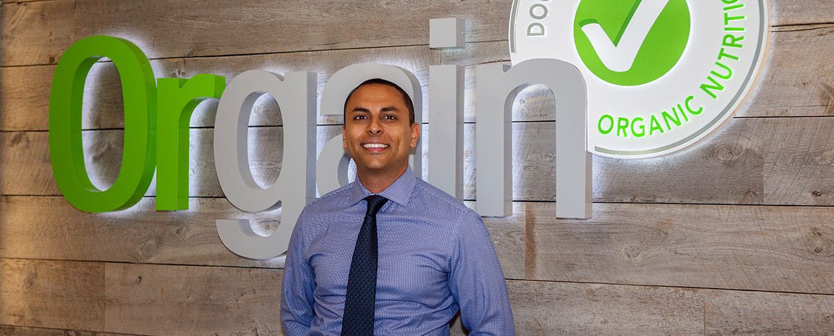 A person stands in front of a large sign that reads "Orgain," with the text "Organic Nutrition" inside a green circle checkmark logo. The background is a wood panel wall, and the person is wearing a blue dress shirt and a dark tie, smiling at the camera.