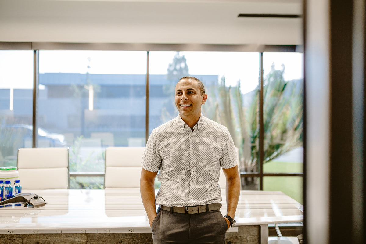 A man is standing and smiling in a modern office with large windows behind him. He is wearing a light-colored, short-sleeve button-up shirt, and gray pants. There is a conference table with white chairs and a few items on it in the background.