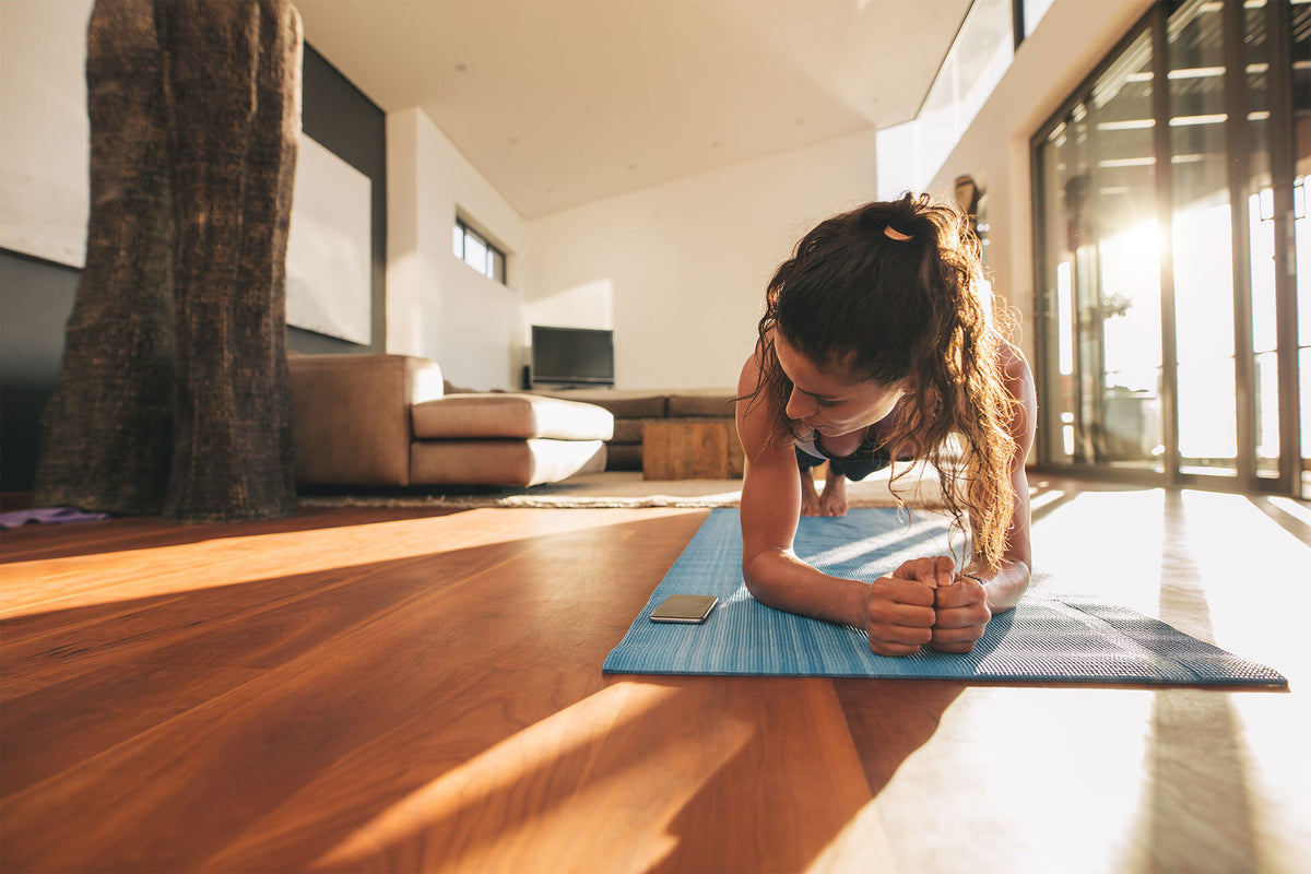 A woman exercises in a plank position on a blue yoga mat in a brightly lit modern living room. She is focused and balanced on her forearms and toes. Nearby, a smartphone rests on the mat. The room features a couch, a tree trunk column, and large windows.