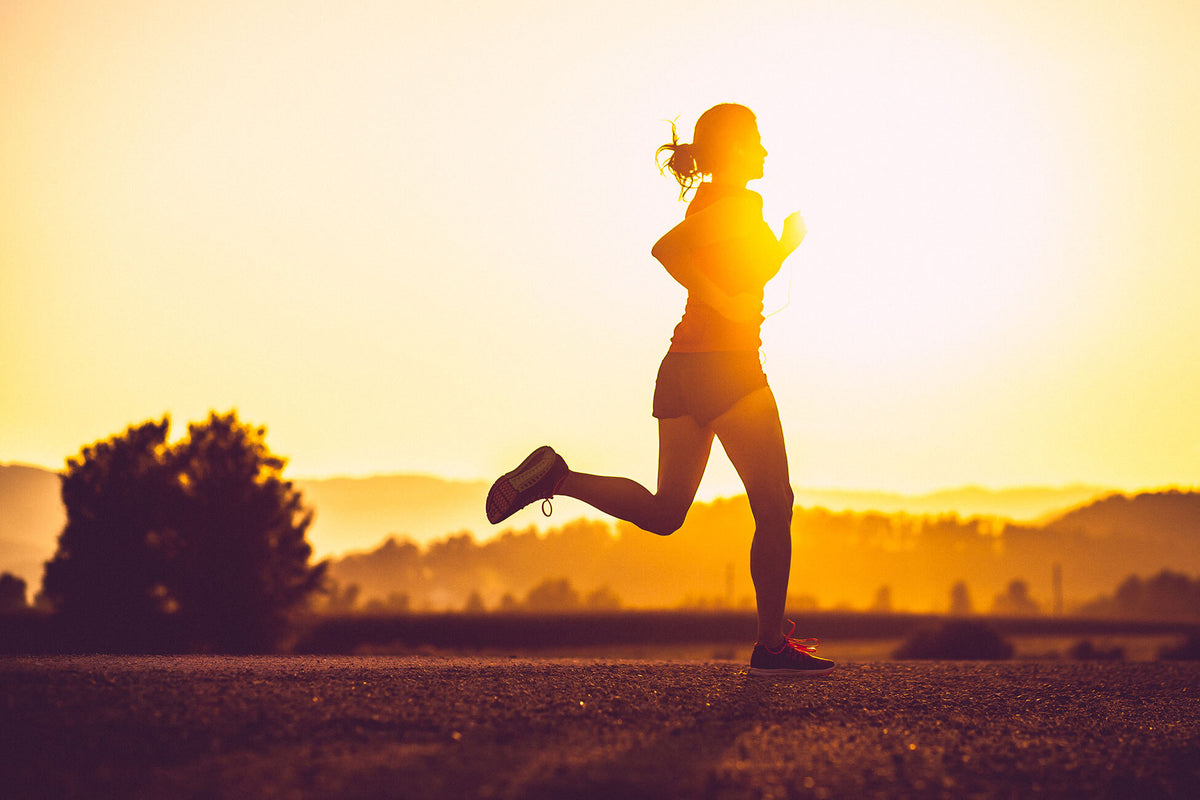 A silhouette of a person running at sunrise or sunset, with a bright orange and yellow sky in the background. The runner is in mid-stride on a flat, outdoor path, with distant hills and trees adding to the serene landscape.