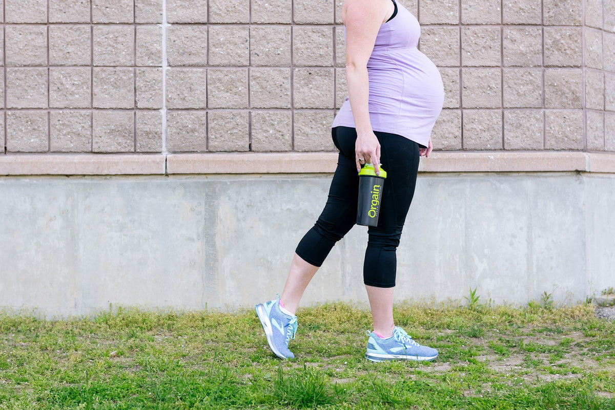 A pregnant person, dressed in a light purple tank top and black leggings, is walking outdoors on a grassy area. They are carrying a green water bottle in their left hand and are wearing blue athletic shoes. The background features a beige brick wall.