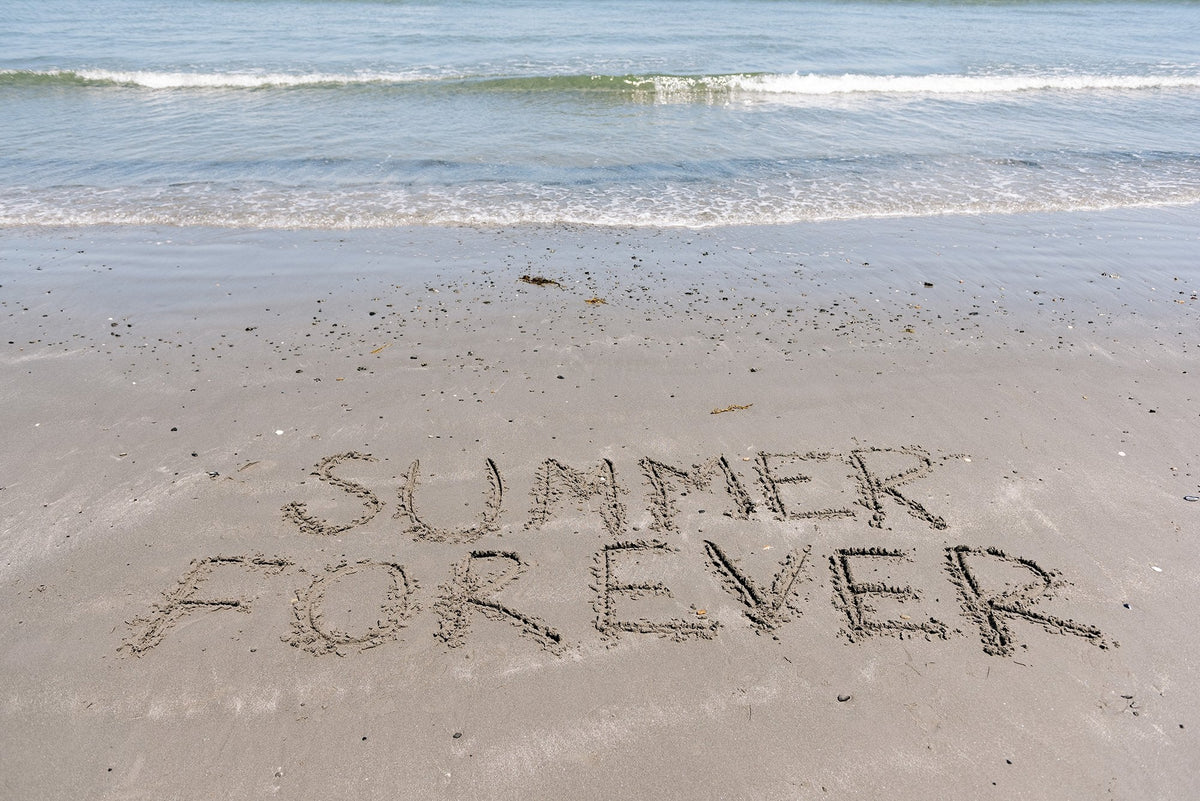 The words "SUMMER FOREVER" are written in large, uppercase letters in the sand on a beach. Behind the writing, gentle waves are lapping at the shore under a clear sky.