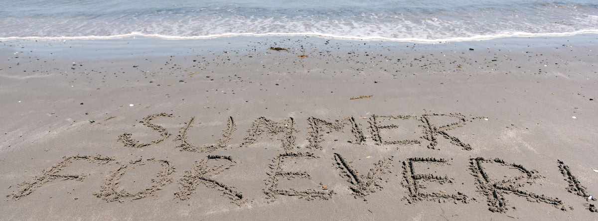 SUMMER FOREVER" is written in the sand on a beach with gentle waves lapping at the shore in the background.