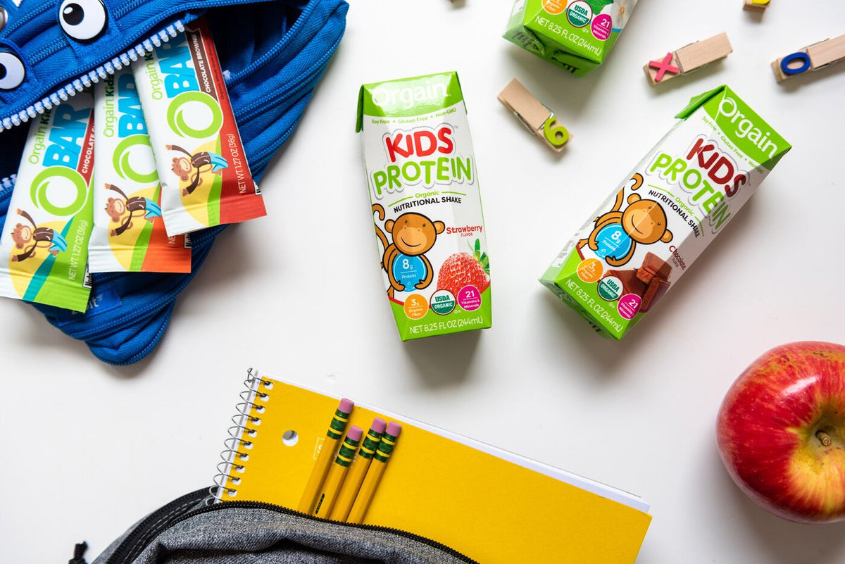 A school backpack with a cartoonish face, an apple, a notebook, colored pencils, alphabet stamps, Orgain Kids Protein shakes, and Orgain protein bars are spread on a white surface. The protein shakes feature a playful monkey graphic on the packaging.