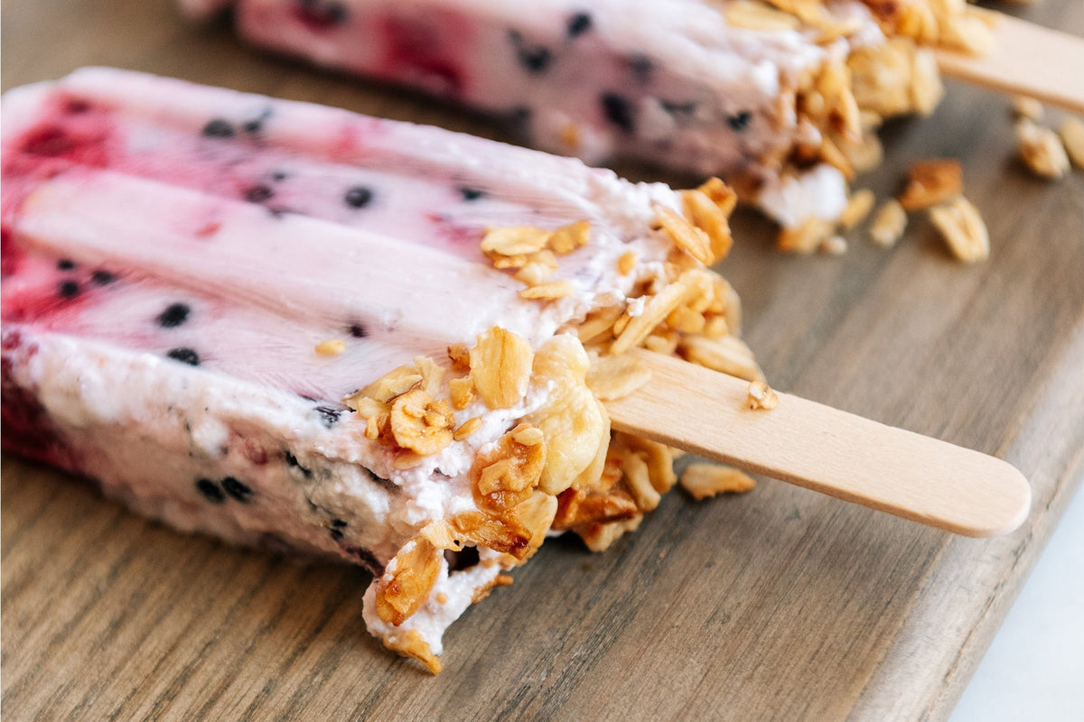 Close-up of two homemade yogurt popsicles with visible blueberries and granola pieces. The popsicles are placed on a wooden surface. The creamy texture and berry swirls are noticeable, complemented by crunchy granola toppings on the sticks.