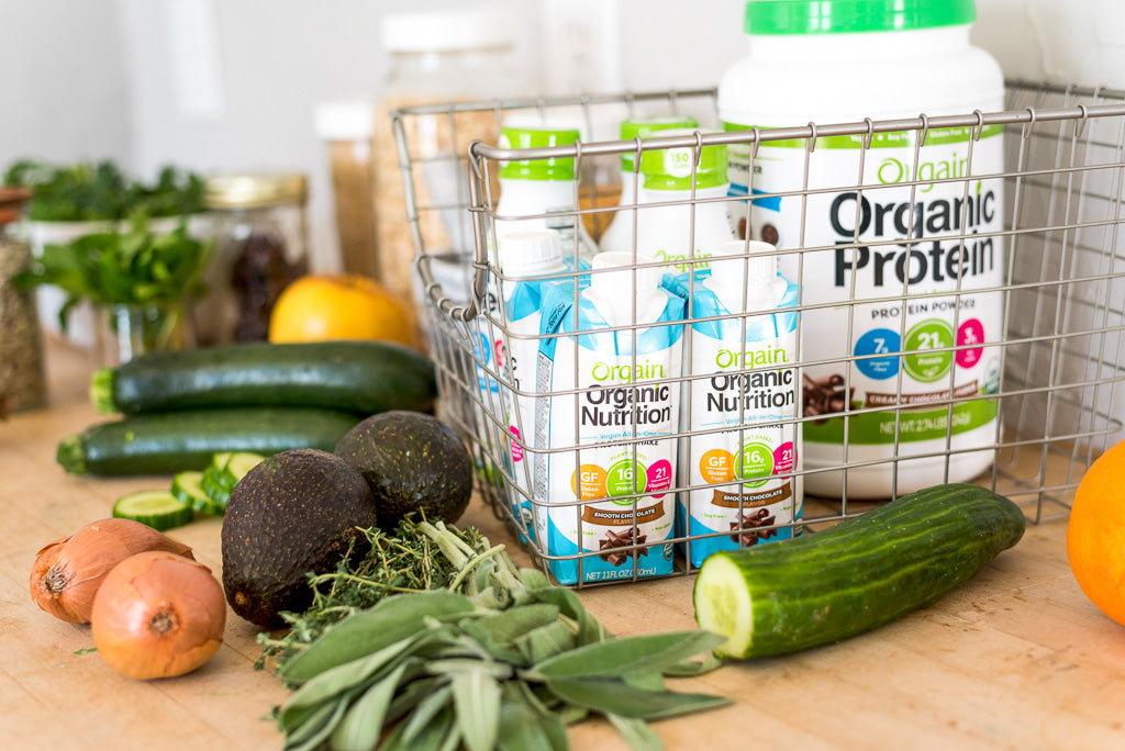 A kitchen counter with fresh vegetables and Orgain protein containers and drinks.