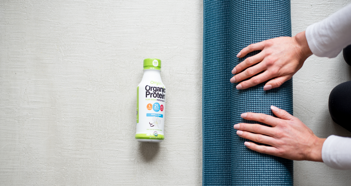 A bottle of Orgain Organic Protein nutritional shake is placed on the floor next to a person‚Äôs hands rolling up a blue yoga mat. The background is a light-colored surface.