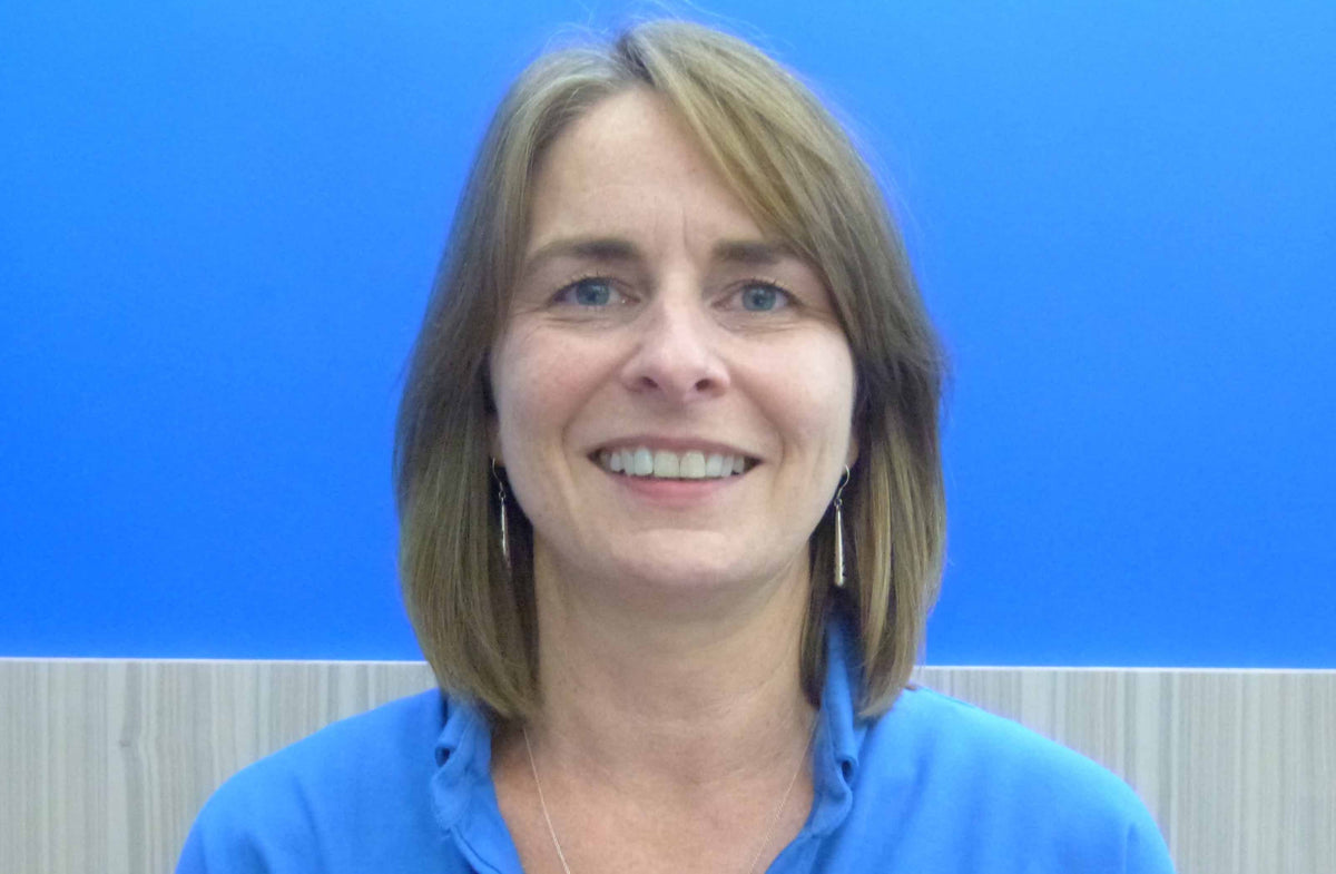 A woman with shoulder-length brown hair and blue eyes is smiling at the camera. She is wearing a blue top and dangling earrings. The background consists of a gradient of blue tones with a light-colored stripe at the bottom.
