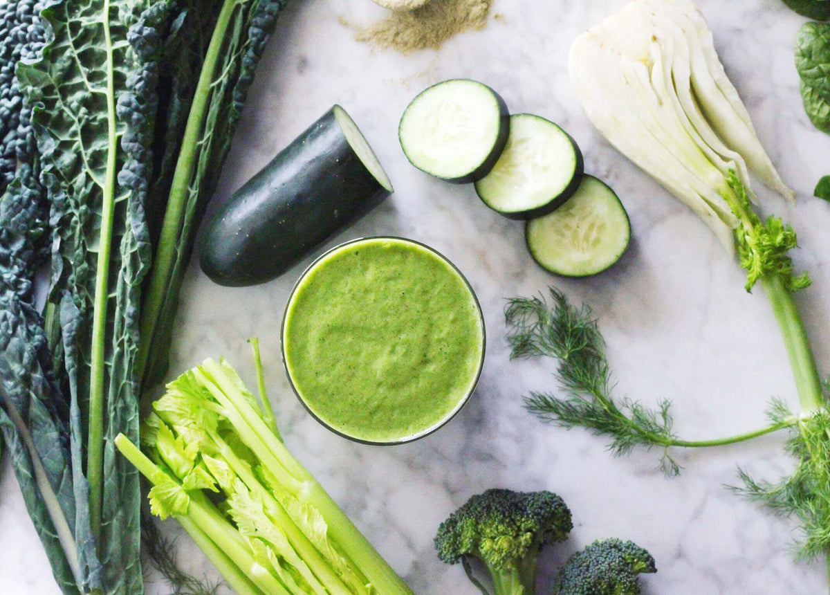 A green smoothie surrounded by green vegetables on the kitchen counter.
