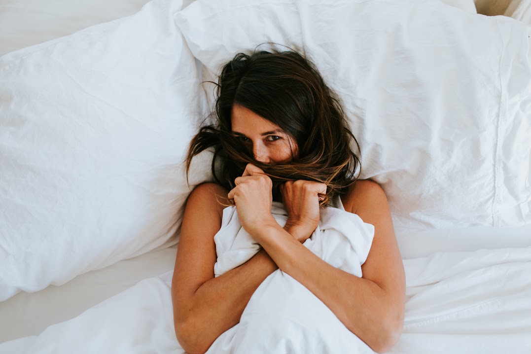 A woman in bed, holding a blanket close to her face, with a playful expression. The bed has white pillows and sheets.
