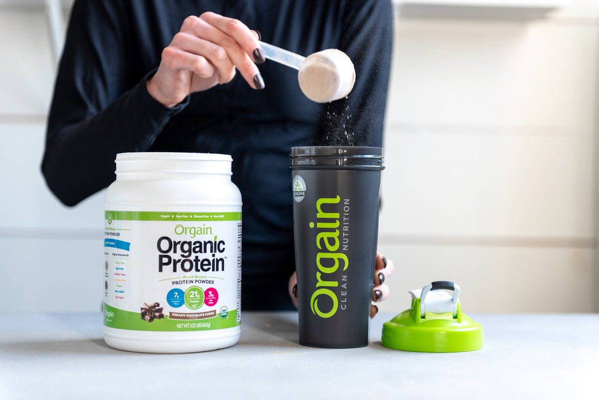 A person is scooping Orgain Organic Protein powder from a white and green container into a black shaker bottle with the Orgain logo. The shaker lid, featuring a green accent, is placed on the table. The person is wearing a dark long-sleeve shirt.