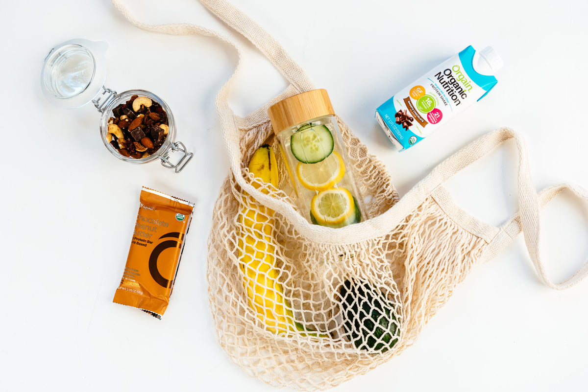 Mesh bag containing lemons, a cucumber, and a glass bottle with cucumber and lemon slices, alongside a snack bar, a jar of mixed nuts, and a carton of plant-based milk on a white surface.