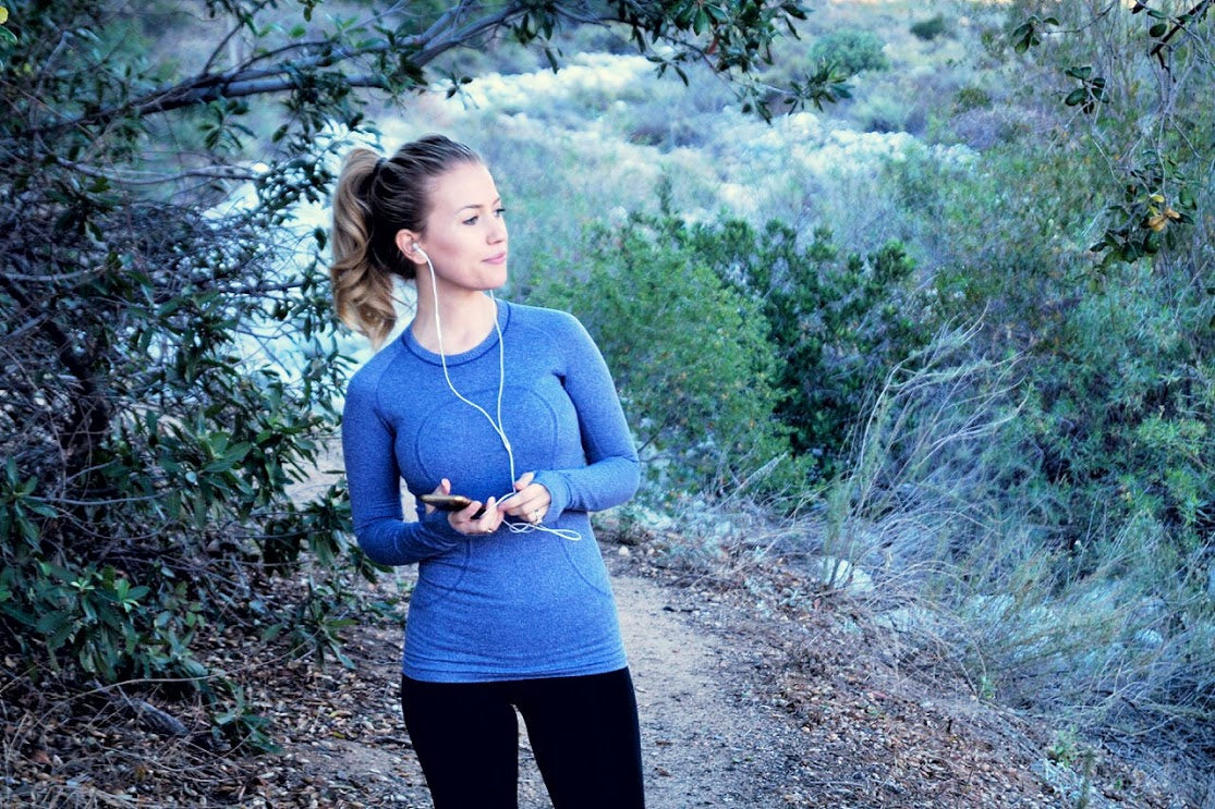 A woman with a ponytail, wearing a blue long-sleeved shirt and black leggings, stands on a forest trail holding a phone and listening to music with earphones. The background features greenery and a mountain landscape.