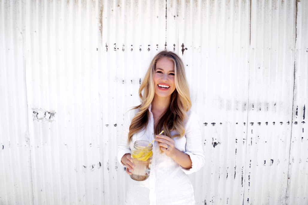 A woman with long blonde hair stands against a white corrugated wall, smiling and holding a glass jar filled with water and lemon slices. She is wearing a white button-up shirt.