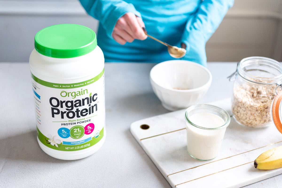 A person in a blue shirt is scooping powder from a container labeled "Orgain Organic Protein" into a white bowl. Nearby are a glass of milk, a jar of oats, and a banana, all placed on a white surface.