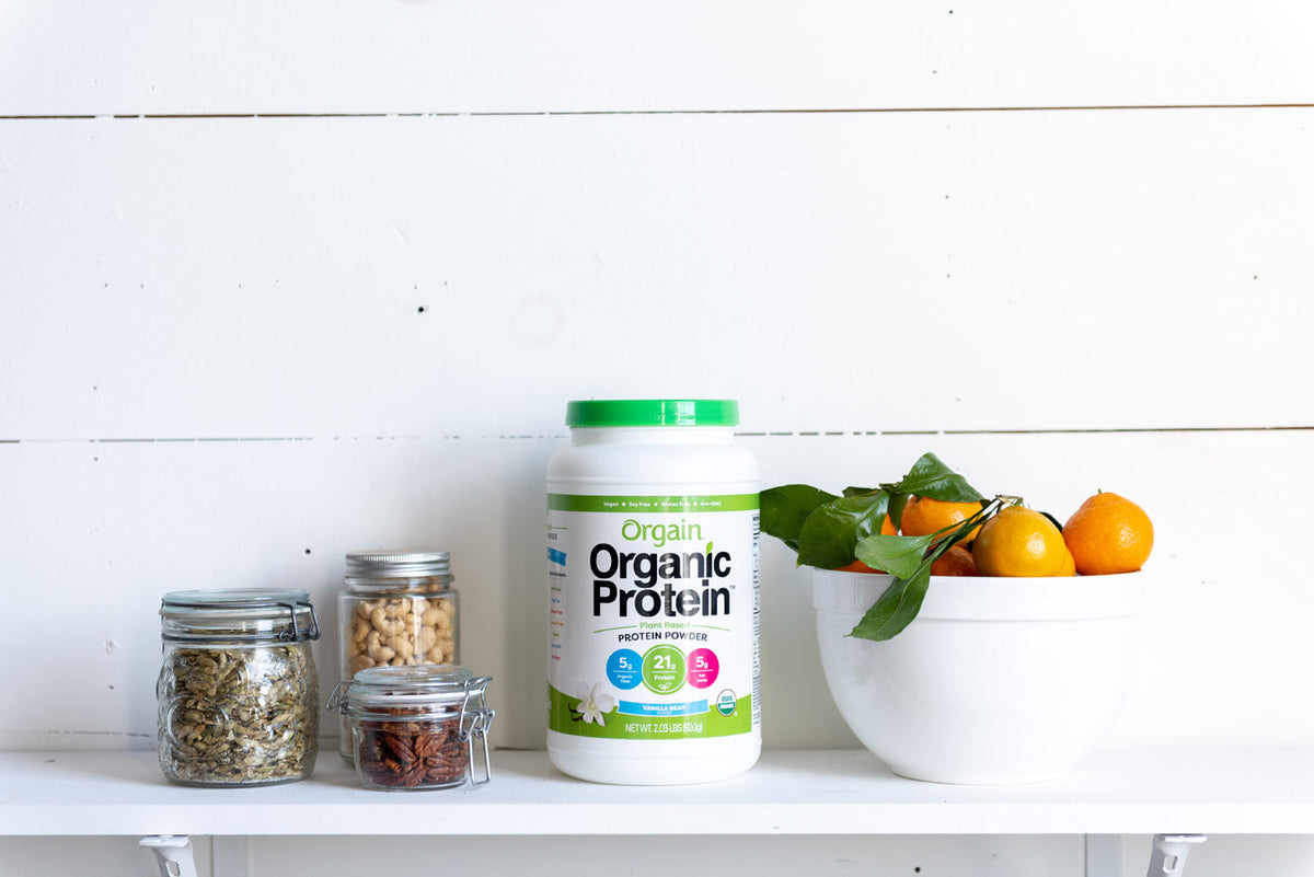 Orgain Organic Protein powder on a white shelf with jars of nuts and seeds and a bowl of oranges.