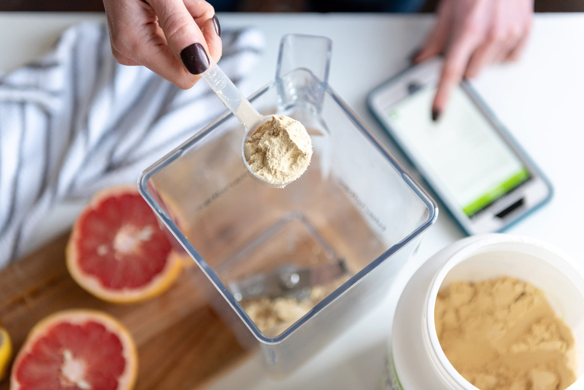 A person adds a scoop of protein powder to a blender. A grapefruit, a knife on a cutting board, and a container of protein powder are visible. The person's other hand is touching a tablet displaying a recipe or nutritional information.