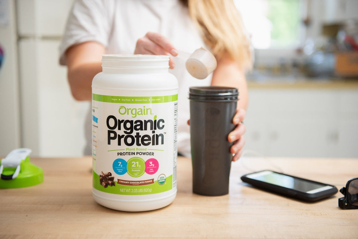 Person scooping Orgain Organic Protein Powder into a black shaker bottle. The protein powder container is prominently displayed on a kitchen counter. Nearby items include a phone, sunglasses, and another lid or shaker component.