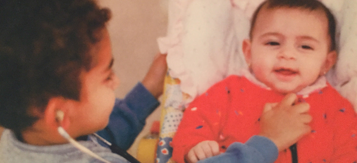 A young boy with curly hair and an earpiece smiles while holding the hand of a baby lying down in a pink outfit. The baby looks content and is also smiling. They appear to be in a cozy indoor setting.