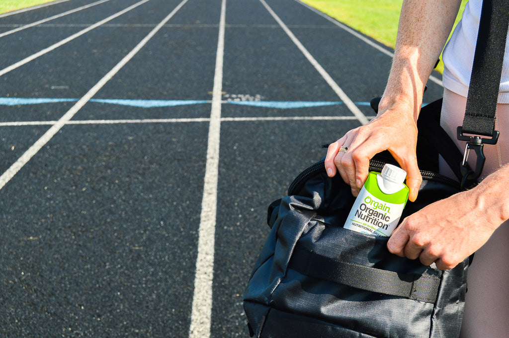 A person on a running track holds a black gym bag and is placing a green and white bottle of Orgain Organic Nutrition into the bag's pocket. The track, marked with white lines, extends into the distance with green grass on either side.