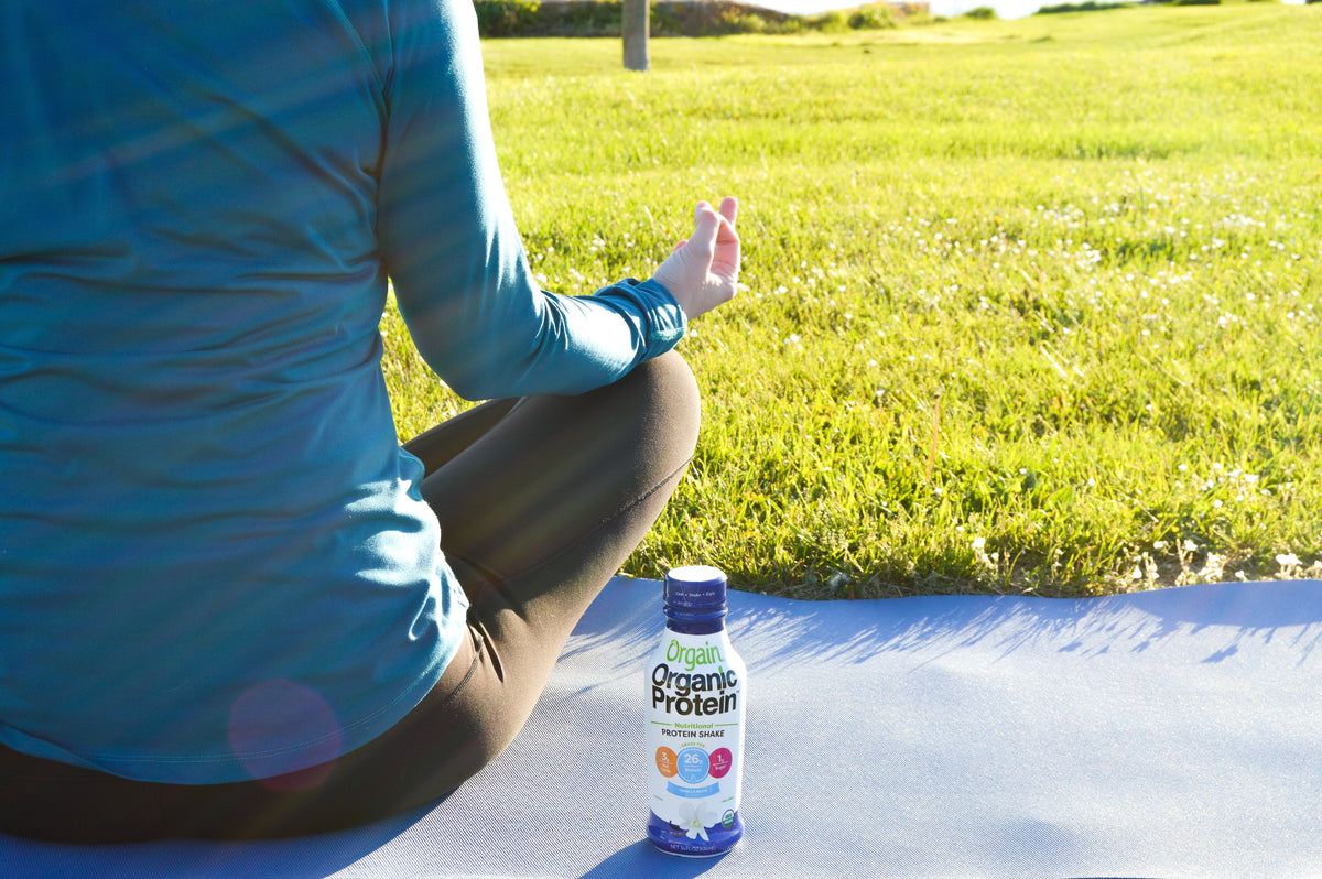 A person sits cross-legged on a yoga mat in a grassy field, meditating with one hand extended and a protein bottle in front.