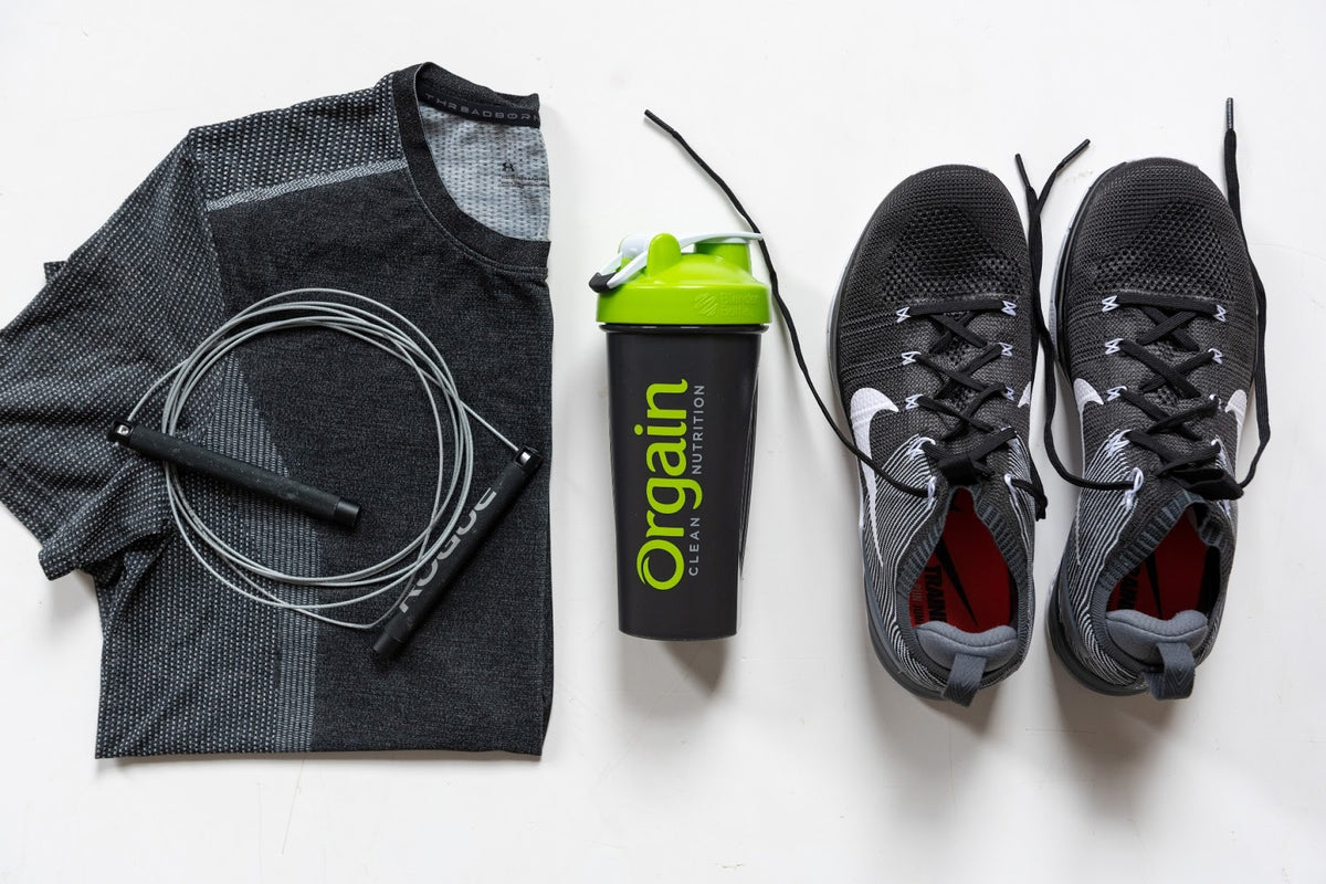 Flat lay photo of workout gear consisting of a grey athletic shirt, a black jump rope with grey handles, a black and green Orgain shaker bottle, and a pair of black and white athletic shoes, all arranged neatly on a white surface.