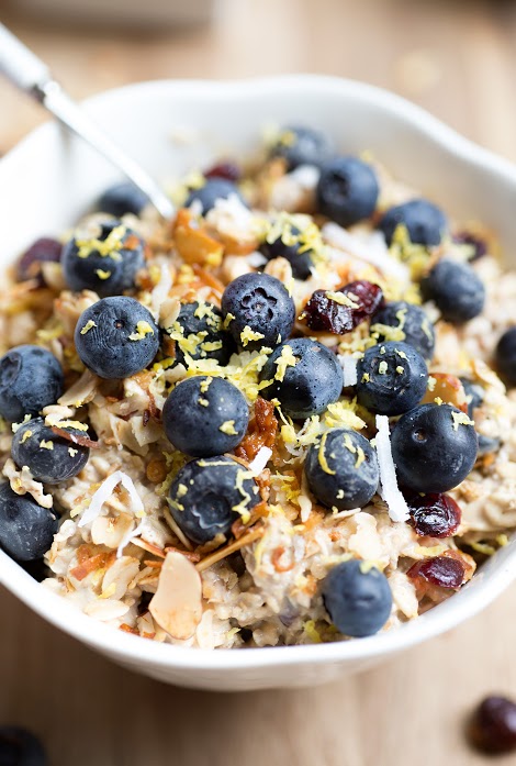 A bowl of oatmeal topped with fresh blueberries, chopped nuts, dried cranberries, and lemon zest. The oatmeal is creamy, and the toppings add vibrant color and texture to the dish. A spoon is placed in the bowl, ready for eating.