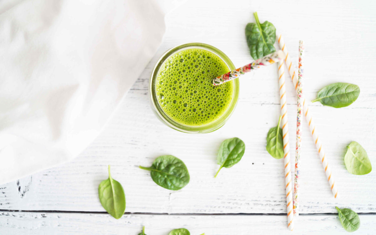 A top view of a green smoothie in a glass with a decorative straw on a white wooden surface. Fresh spinach leaves are scattered around the smoothie, along with a few additional straws. A white fabric is partially visible in the upper left corner.