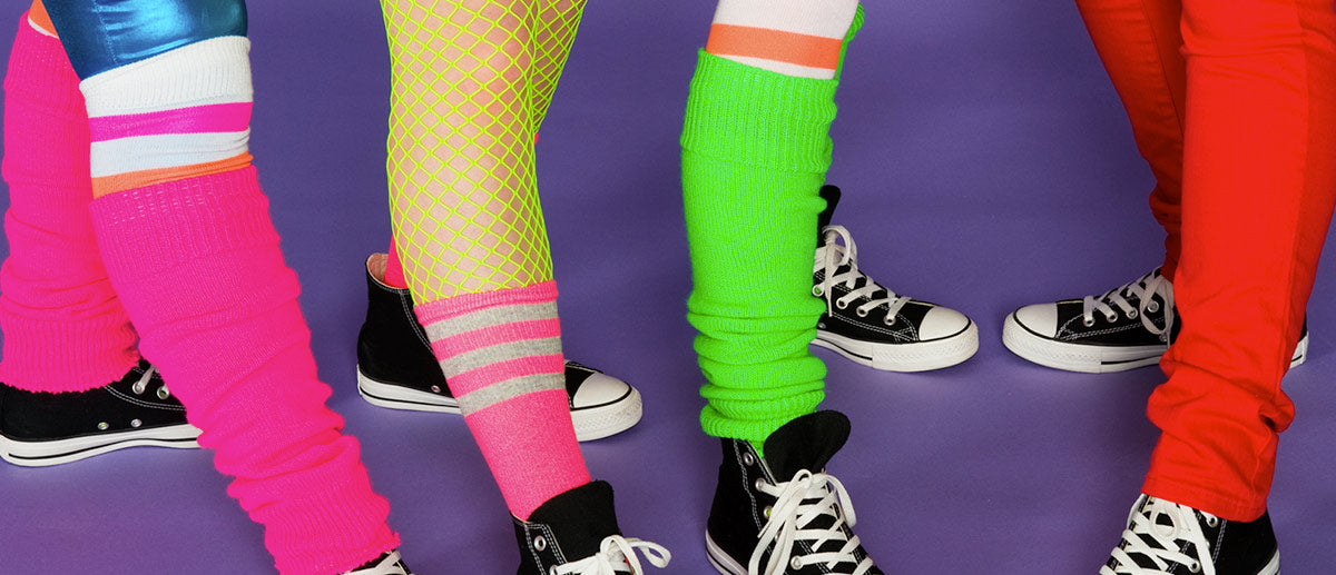 Five people are standing on a purple floor wearing 80s-style colorful leg warmers, neon fishnet stockings, and sneakers. The vibrant attire includes hot pink, green, and red leg warmers over various tights and leggings, creating a retro fashion vibe.