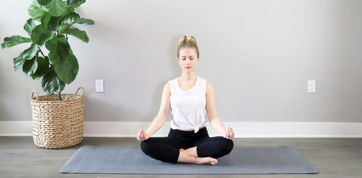 A woman sits cross-legged on a yoga mat, meditating with palms facing upward, in a room with a large potted plant.