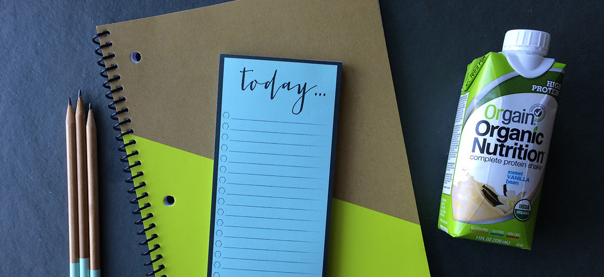A spiral-bound notebook, a daily to-do list with "today..." written at the top, three pencils, and a carton of Orgain Organic Nutrition complete protein shake in vanilla flavor are arranged on a dark surface.