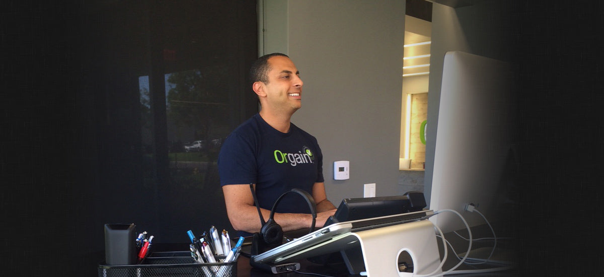 A person in an "Orgain" t-shirt smiles at a computer screen on a desk with office supplies and a headset.