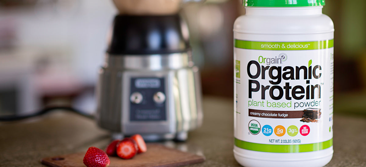 A container of Orgain Organic Protein (creamy chocolate fudge flavor) on a kitchen counter, with a blender and a cutting board with sliced strawberries in the background.