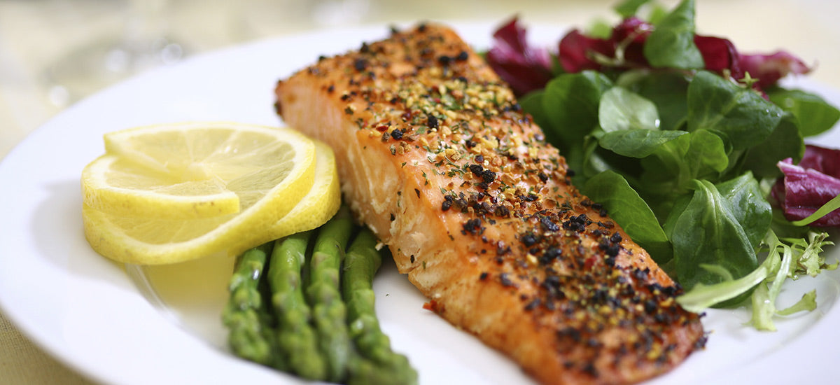 A plate of seasoned, grilled salmon garnished with herbs, served with a side of fresh asparagus, mixed greens, and lemon slices.