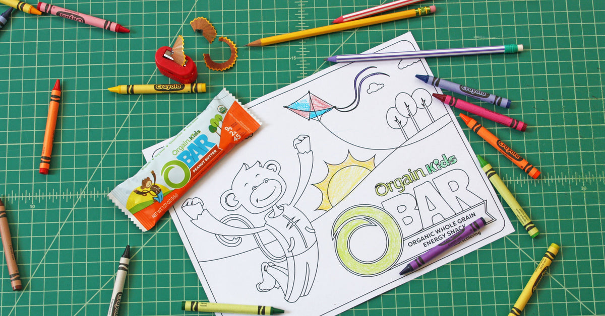 A children's coloring sheet featuring a monkey, a sun, and a kite is surrounded by Crayola crayons on a green cutting mat. A partially eaten organic kids' snack bar and its wrapper are placed on the left side of the coloring sheet.